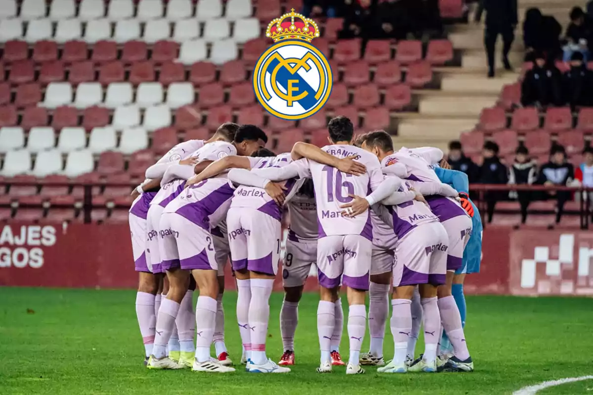 A group of soccer players in purple uniforms hug in a circle in a stadium with empty stands and the Real Madrid crest above them.