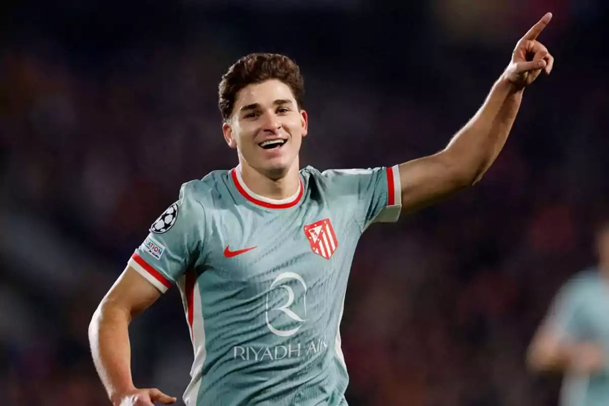Un jugador de fútbol celebrando un gol con el brazo levantado y sonriendo, vistiendo un uniforme gris con detalles rojos y un escudo en el pecho.