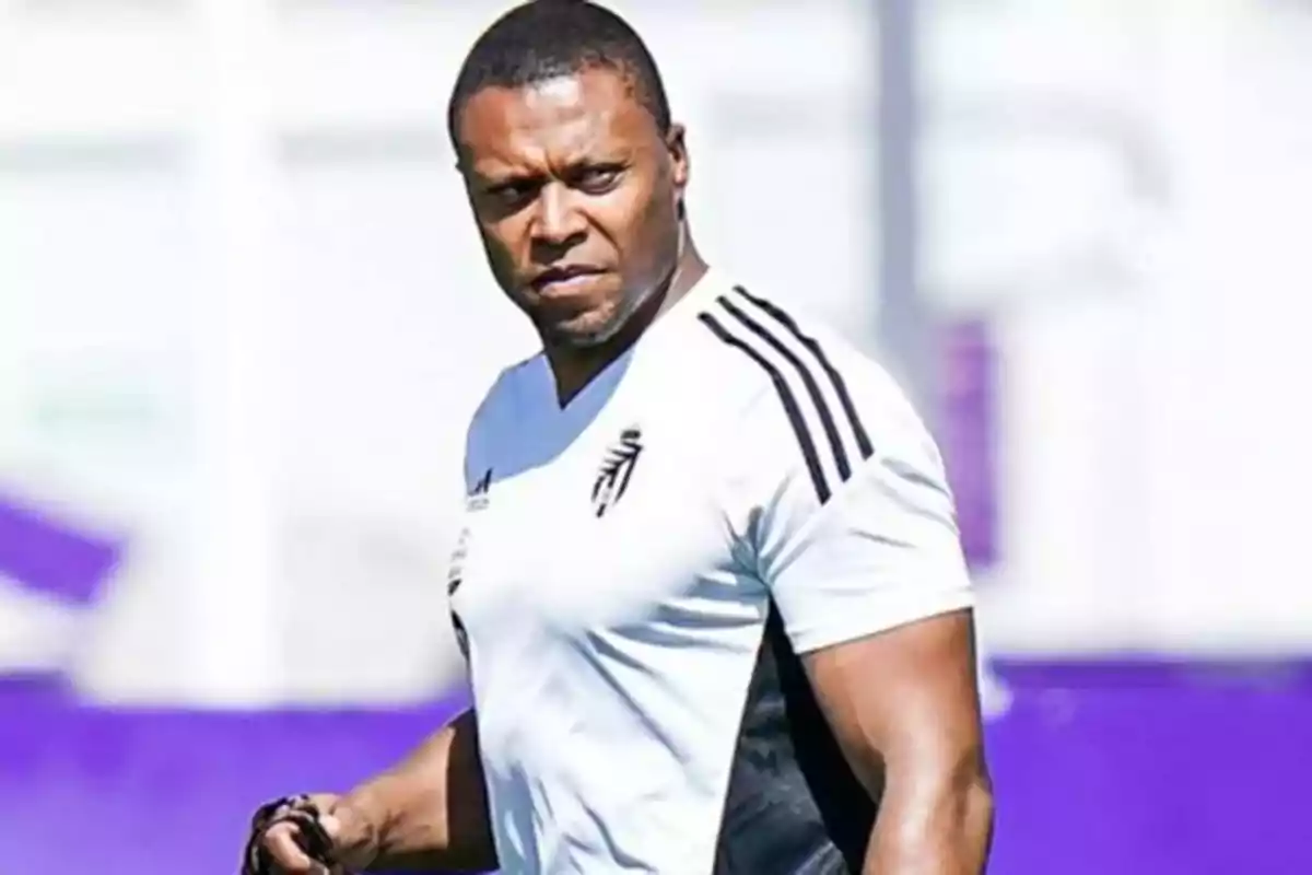 Un hombre con camiseta deportiva blanca y detalles negros en un campo de entrenamiento. Un hombre con camiseta deportiva blanca y detalles negros en un campo de entrenamiento.
