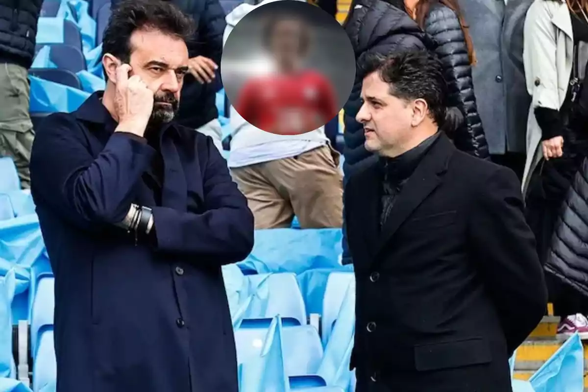 Two men standing in a stadium, one talking on the phone and the other looking at him, with blue seats and people in the background.