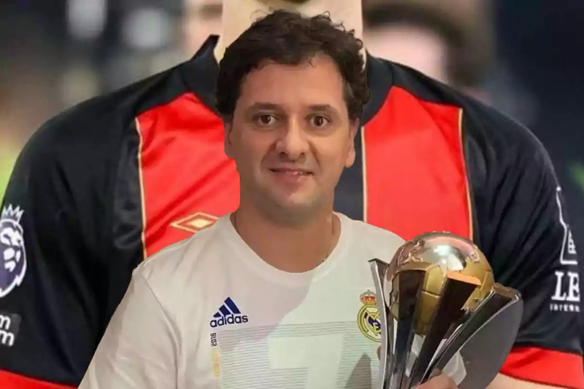 Un hombre con una camiseta blanca del Real Madrid sostiene un trofeo frente a una imagen de fondo de una camiseta de fútbol roja y negra.