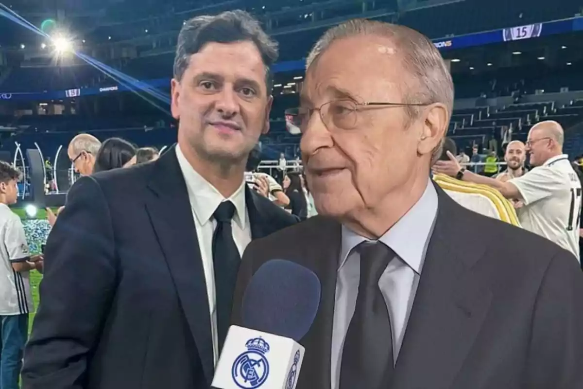 Two men in dark suits at a soccer stadium, one of them holding a microphone with a sports club logo.