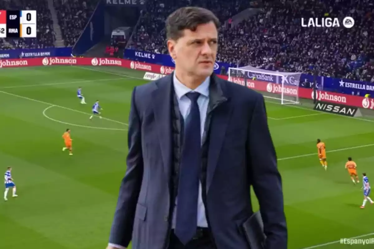 Un hombre con traje está de pie frente a un campo de fútbol durante un partido de LaLiga.