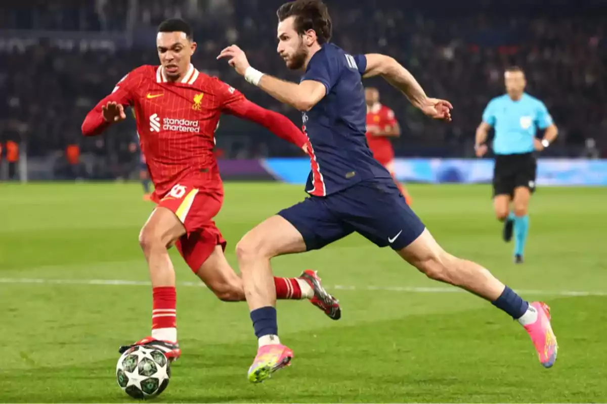 Two players compete for the ball in a match, one wearing red and the other blue, while a referee watches from the background.