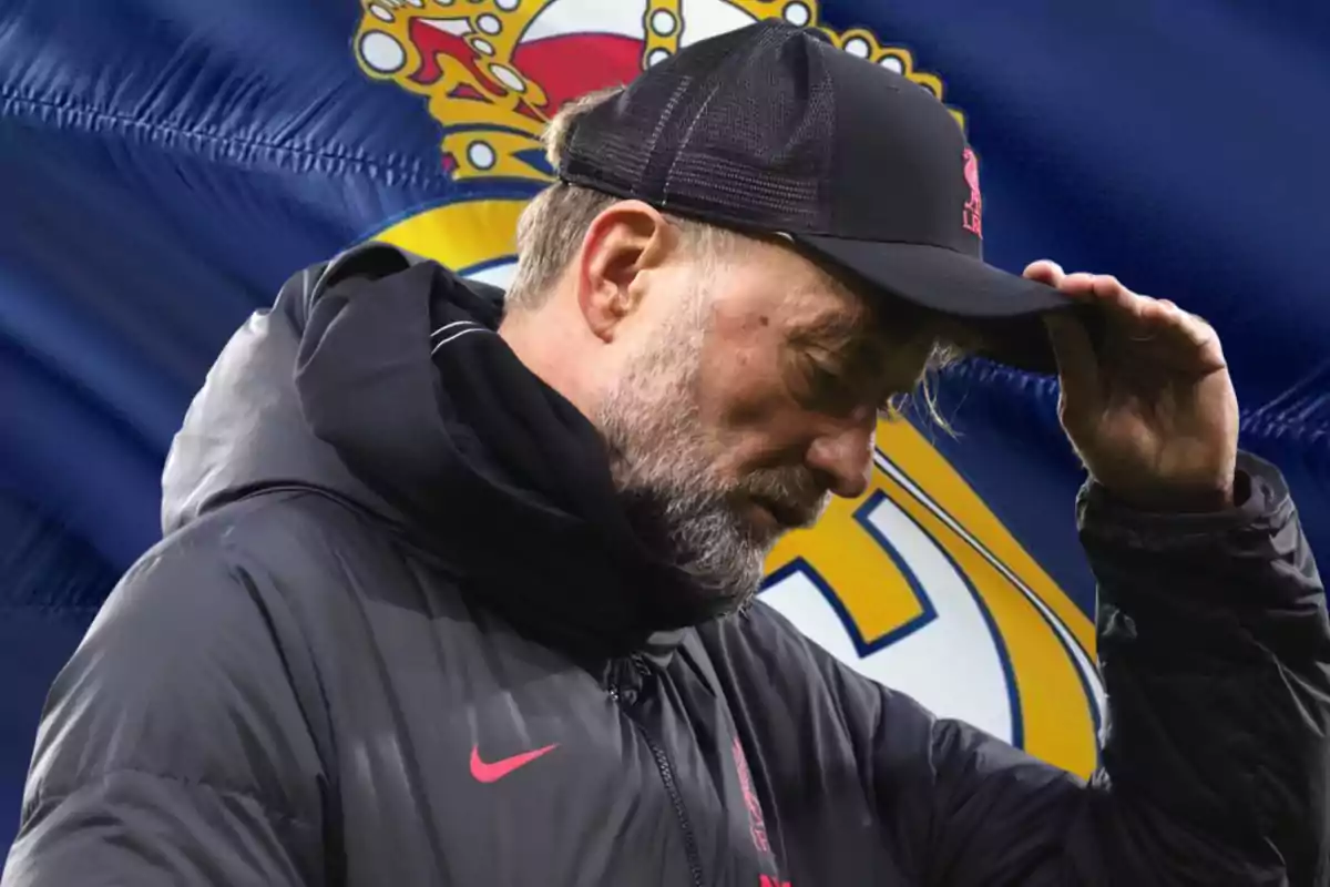 A man in a cap and dark jacket tilts his head in front of a background with a soccer team's emblem.