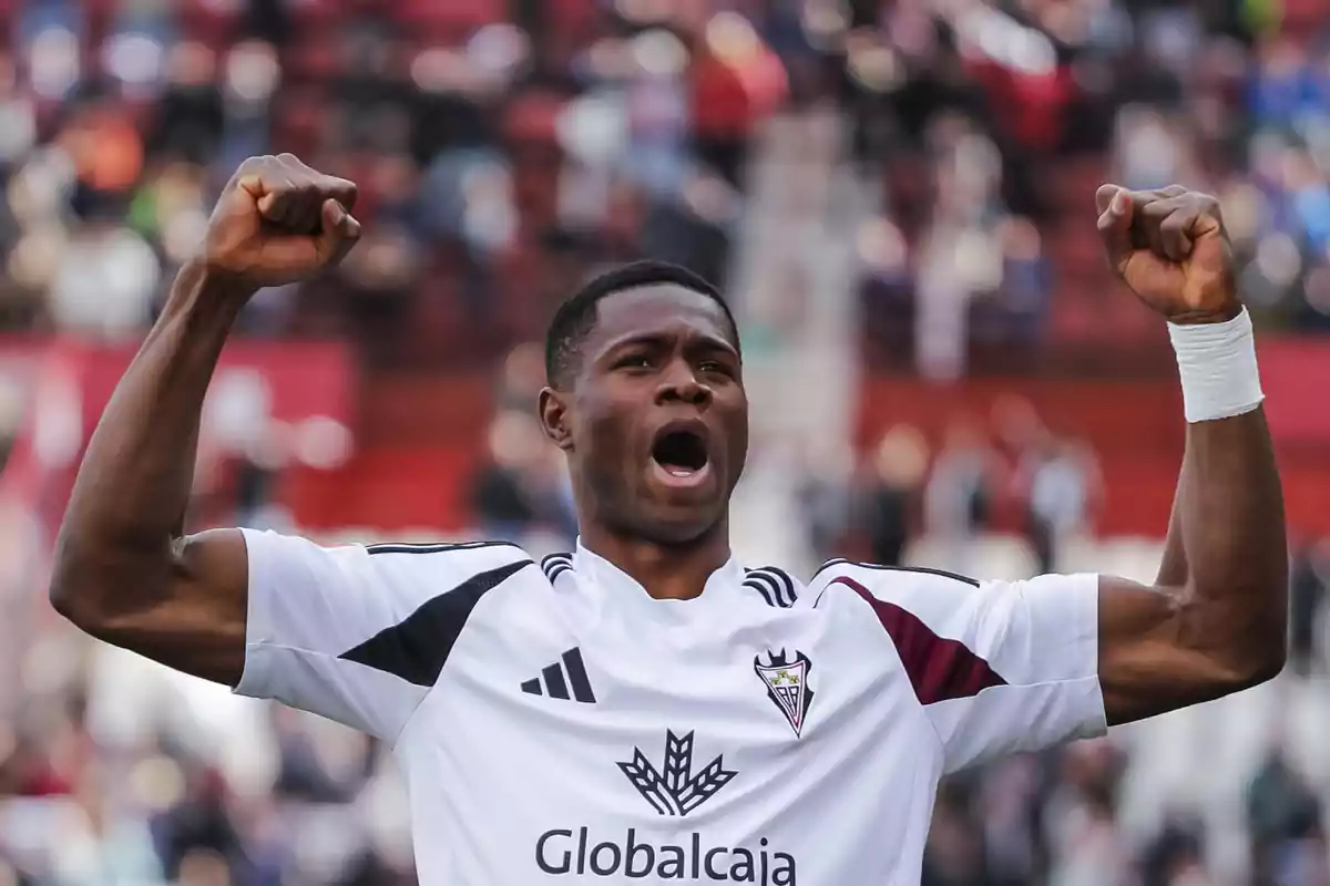 Soccer player celebrating with arms raised, wearing the white Albacete Balompié uniform in a stadium full of fans Soccer player celebrating with arms raised, wearing the white Albacete Balompié uniform in a stadium full of fans