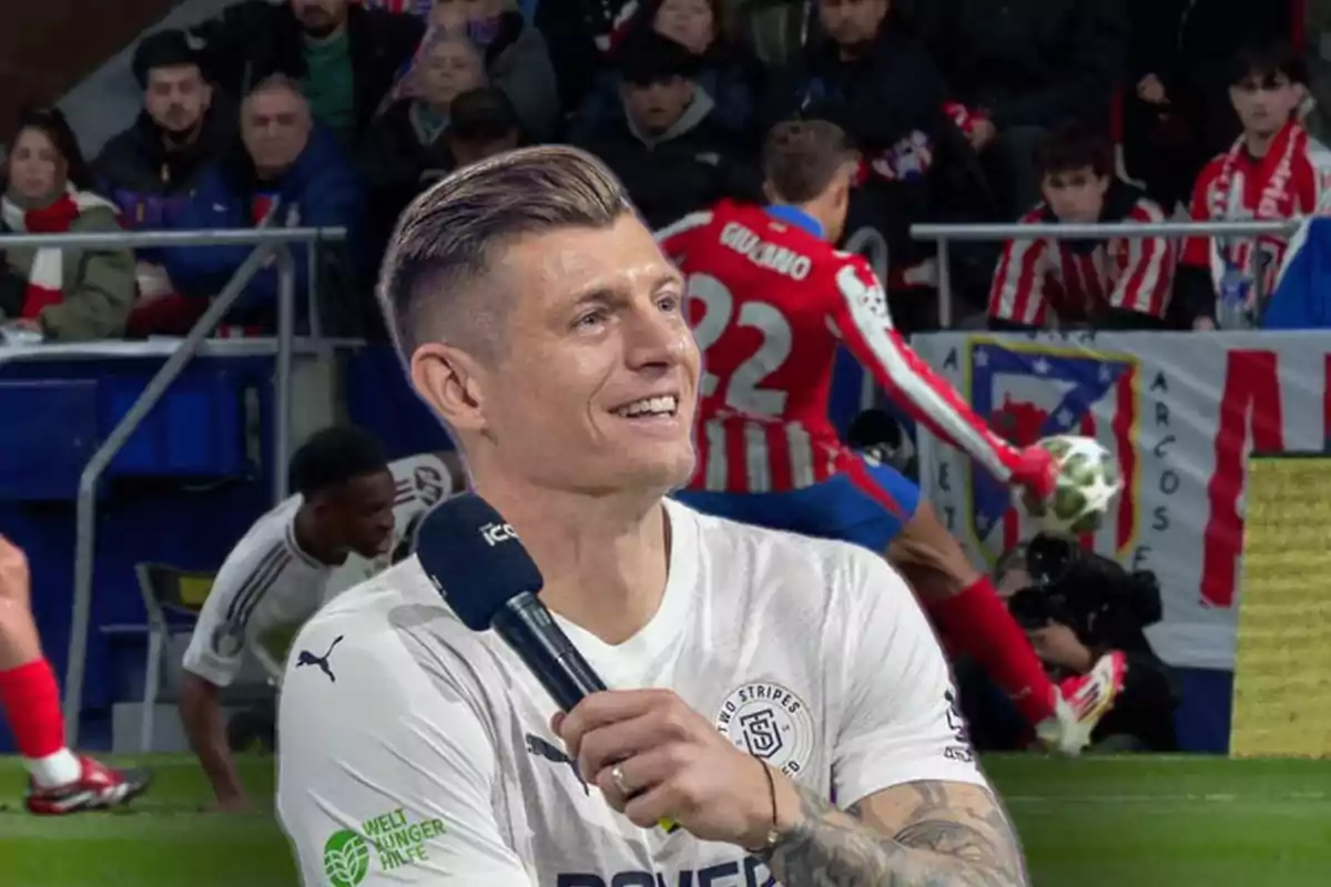 Un hombre sonriente sostiene un micrófono mientras al fondo se observa a un jugador de fútbol en acción en un estadio lleno de espectadores.