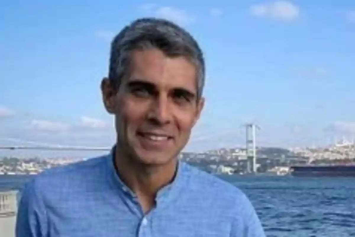 Gray-haired man in a light blue jersey smiling in front of a body of water with a bridge and a city in the background on a sunny day Gray-haired man in a light blue jersey smiling in front of a body of water with a bridge and a city in the background on a sunny day