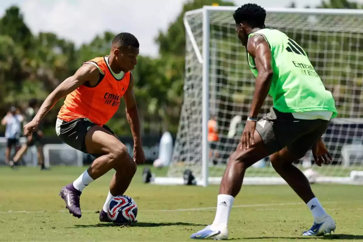 Two players are training on a soccer field; one wearing an orange bib controls the ball while the other, in a green bib, defends against him in front of a goal. Two players are training on a soccer field; one wearing an orange bib controls the ball while the other, in a green bib, defends against him in front of a goal.