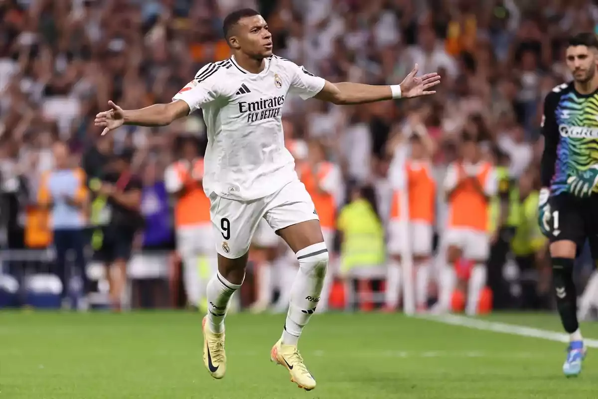 Soccer player celebrating a goal on the field with his arms outstretched while the crowd applauds in the background.