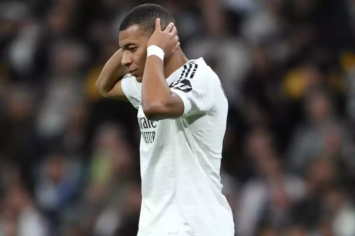 Un jugador de fútbol con uniforme blanco se lleva las manos a la cabeza en un estadio lleno de espectadores.