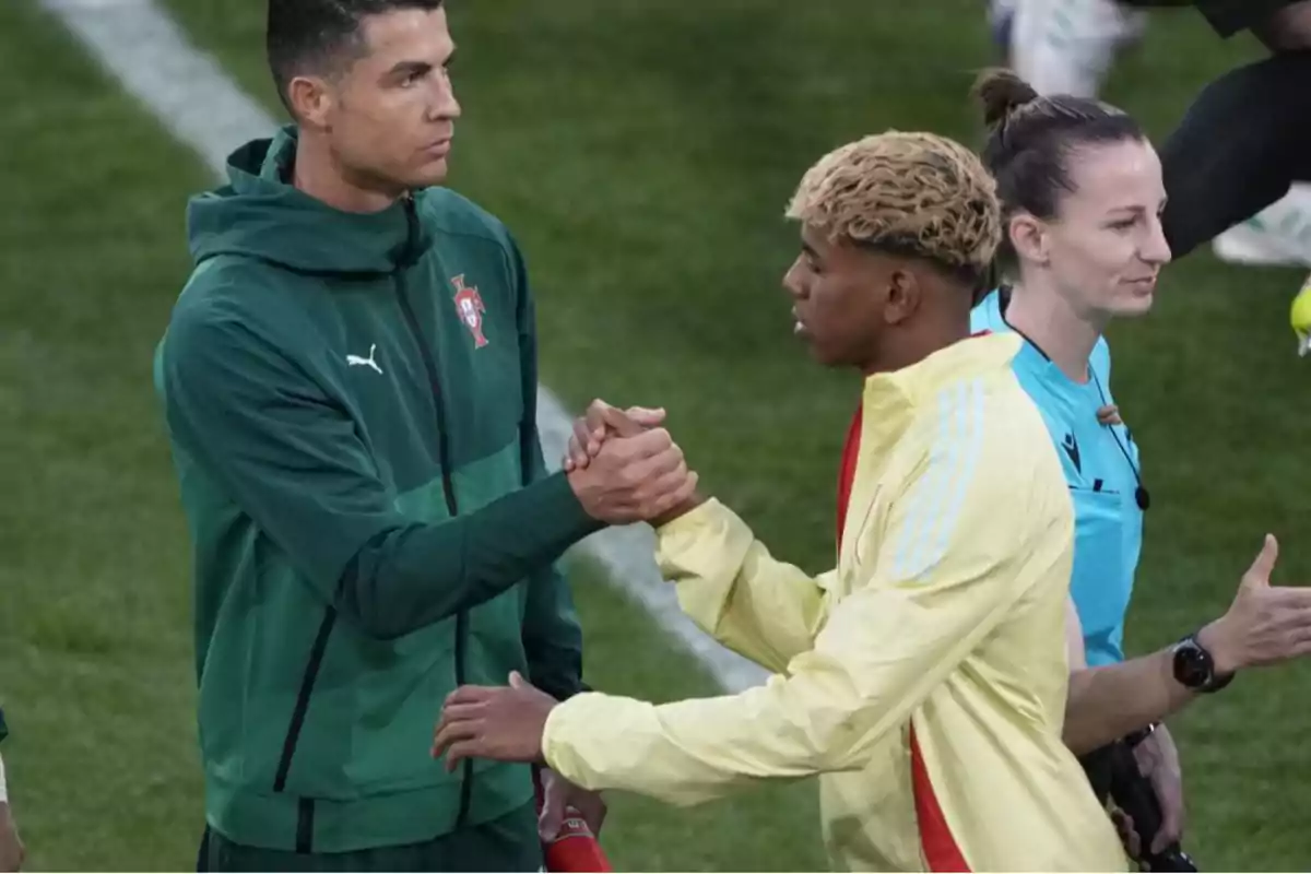 Dos futbolistas se saludan con un apretón de manos en el campo antes de un partido mientras una árbitra camina cerca de ellos Dos futbolistas se saludan con un apretón de manos en el campo antes de un partido mientras una árbitra camina cerca de ellos