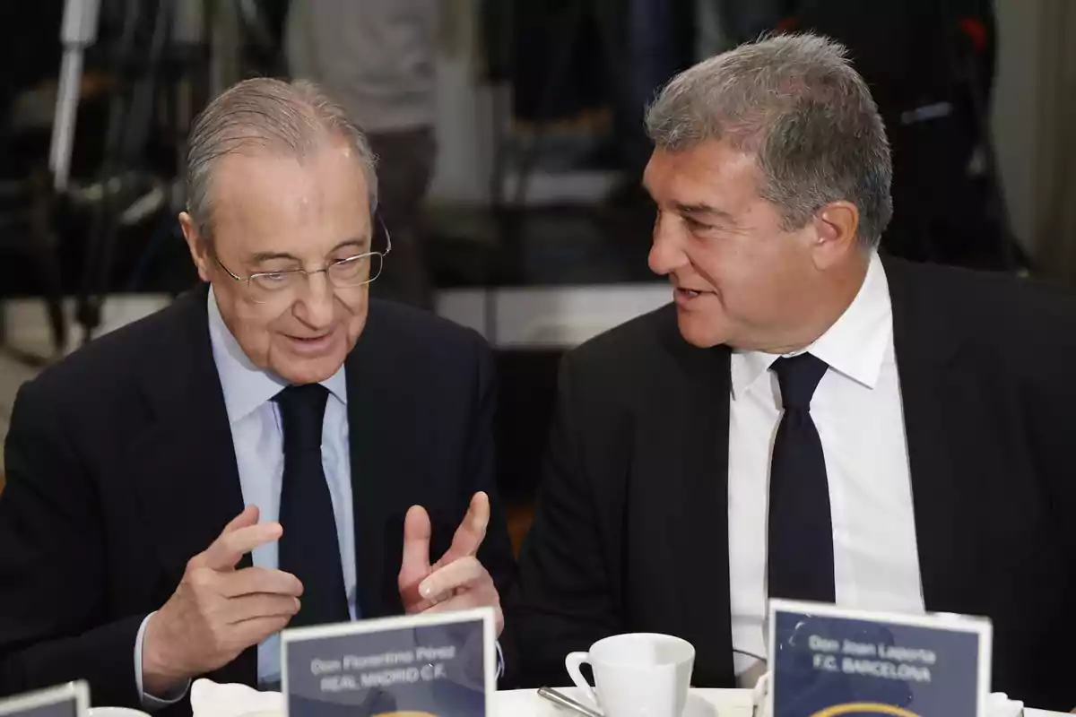 Dos hombres de traje conversan en una mesa con tazas de café frente a ellos. Dos hombres de traje conversan en una mesa con tazas de café frente a ellos.