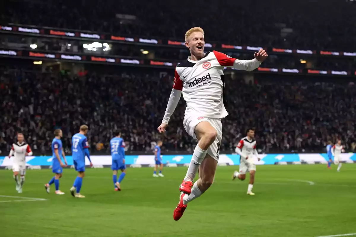 Futbolista de Eintracht Frankfurt celebrando un gol saltando en el aire durante un partido en un estadio lleno de aficionados Futbolista de Eintracht Frankfurt celebrando un gol saltando en el aire durante un partido en un estadio lleno de aficionados
