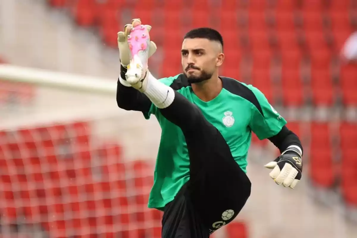 Un futbolista con uniforme verde y guantes de portero estira su pierna durante un entrenamiento en un estadio con asientos rojos. Un futbolista con uniforme verde y guantes de portero estira su pierna durante un entrenamiento en un estadio con asientos rojos.