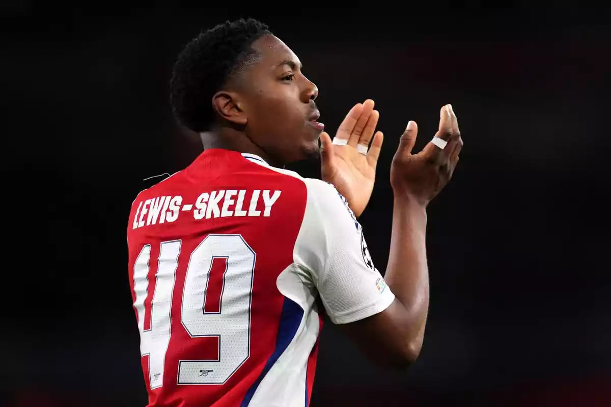 Soccer player in Arsenal’s red and white uniform applauding during a match, with the name Lewis-Skelly and the number 49 on the jersey Soccer player in Arsenal’s red and white uniform applauding during a match, with the name Lewis-Skelly and the number 49 on the jersey