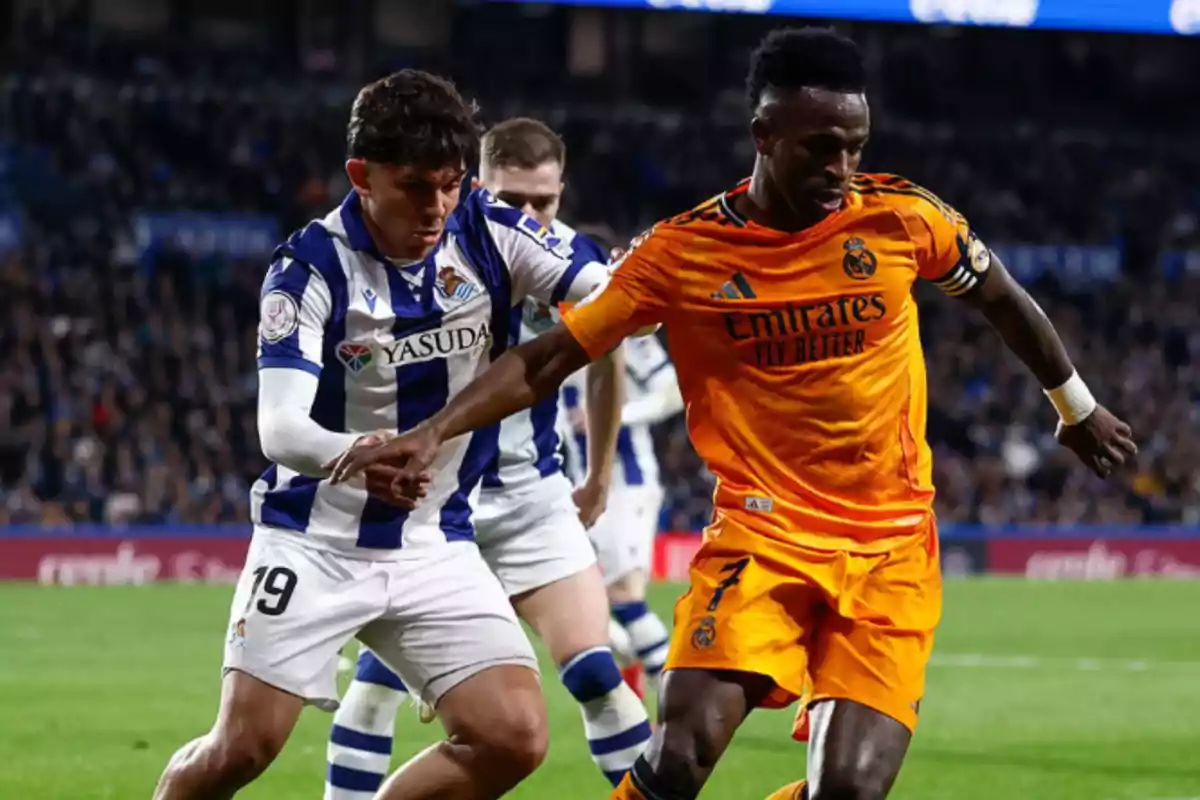 Two players compete for the ball during a match, one wearing a blue and white striped uniform and the other an orange uniform.