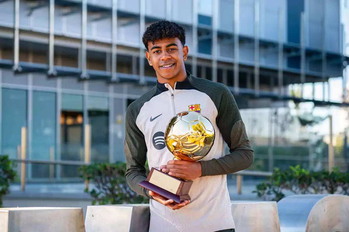 Un joven con uniforme deportivo sostiene un trofeo dorado frente a un edificio moderno.