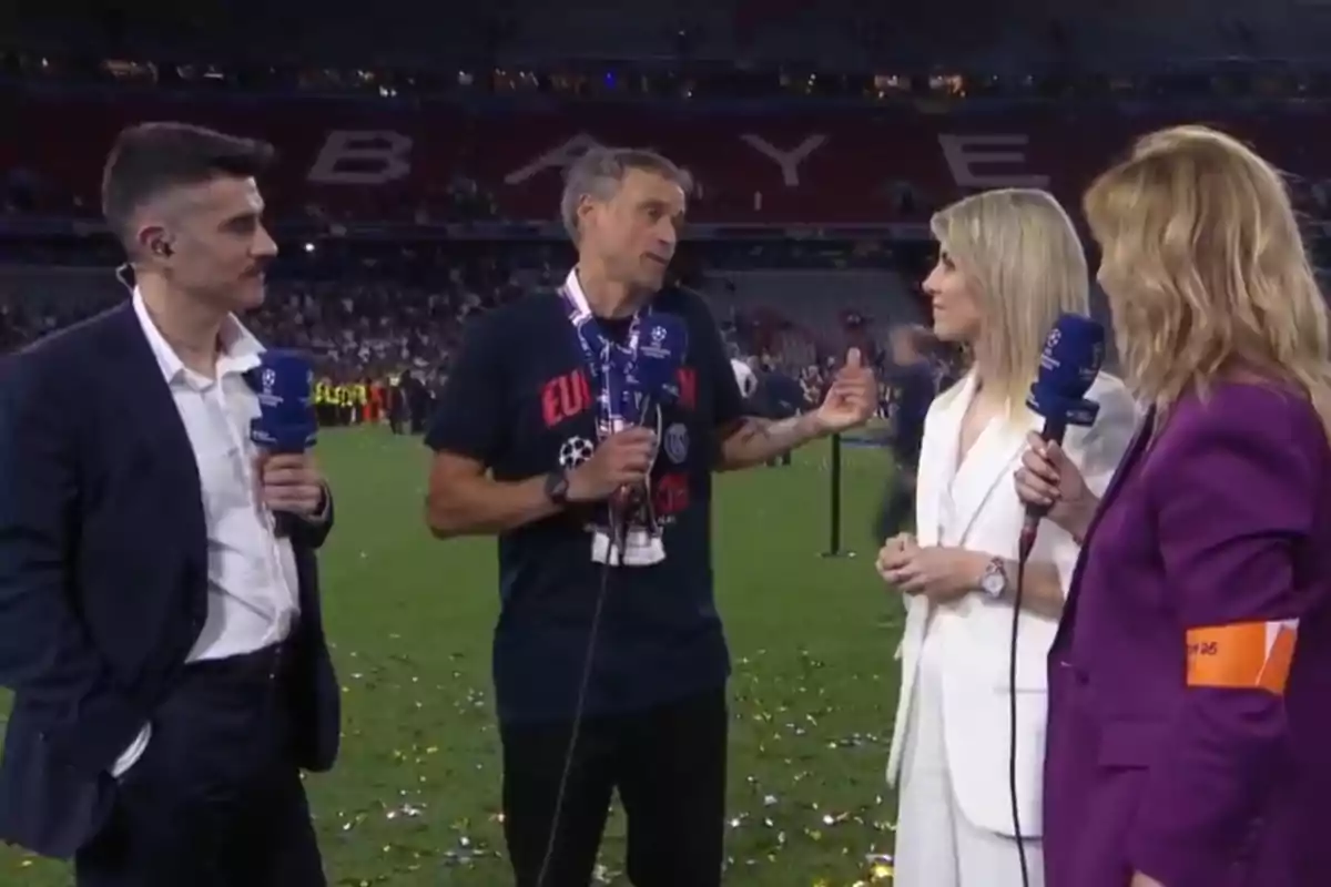 Four people are standing in a football stadium during an on-field interview, holding microphones and talking, with an audience and confetti on the grass.