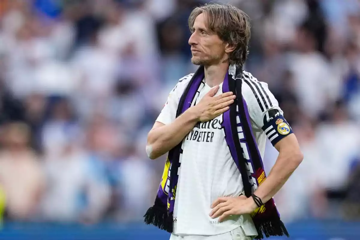 Soccer player in a white uniform and a black and purple scarf places his hand on his chest in a stadium full of fans Soccer player in a white uniform and a black and purple scarf places his hand on his chest in a stadium full of fans
