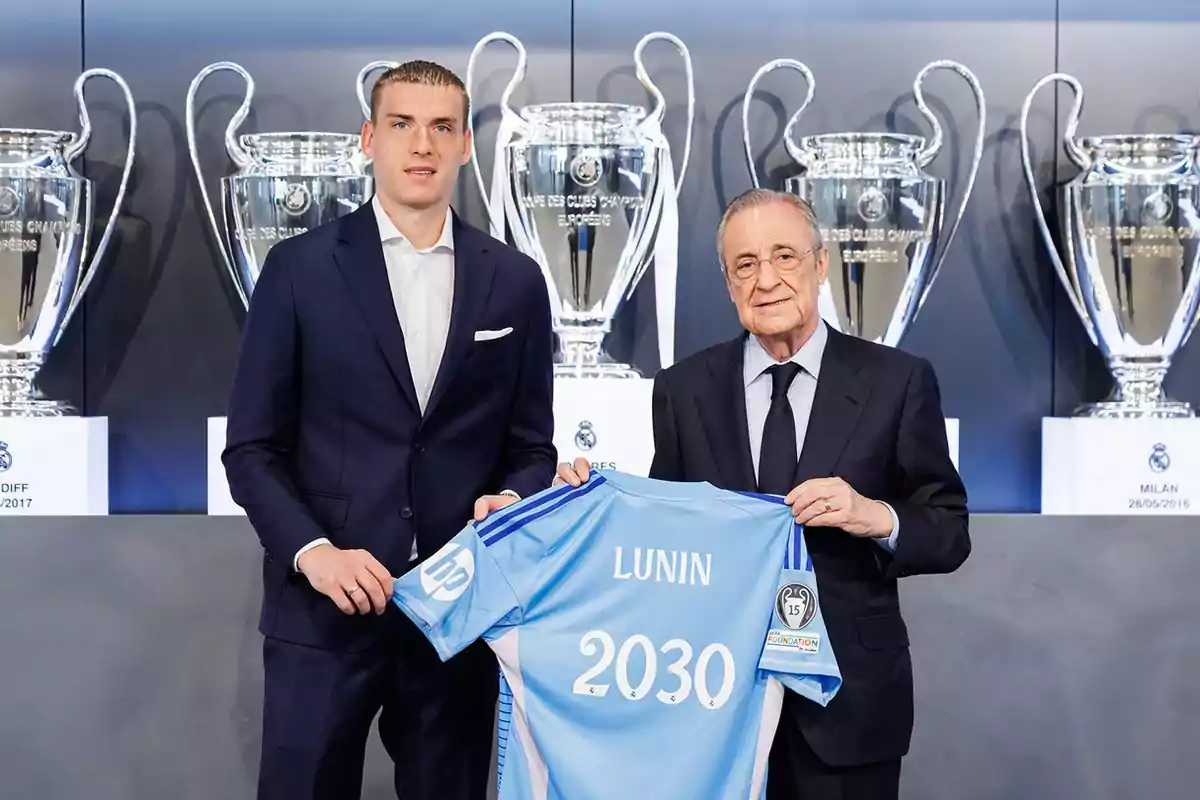Two men in suits pose holding a blue soccer jersey with the name Lunin and the number 2030 in front of several large Champions League trophies. Two men in suits pose holding a blue soccer jersey with the name Lunin and the number 2030 in front of several large Champions League trophies.
