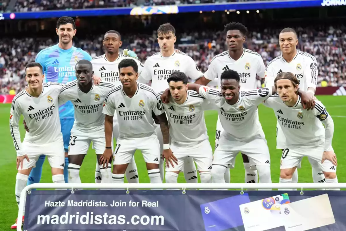 A soccer team posing on the field before a match, all wearing white uniforms with a sponsor's logo and the club's crest, with a crowd in the stands behind them and a promotional banner at the bottom.