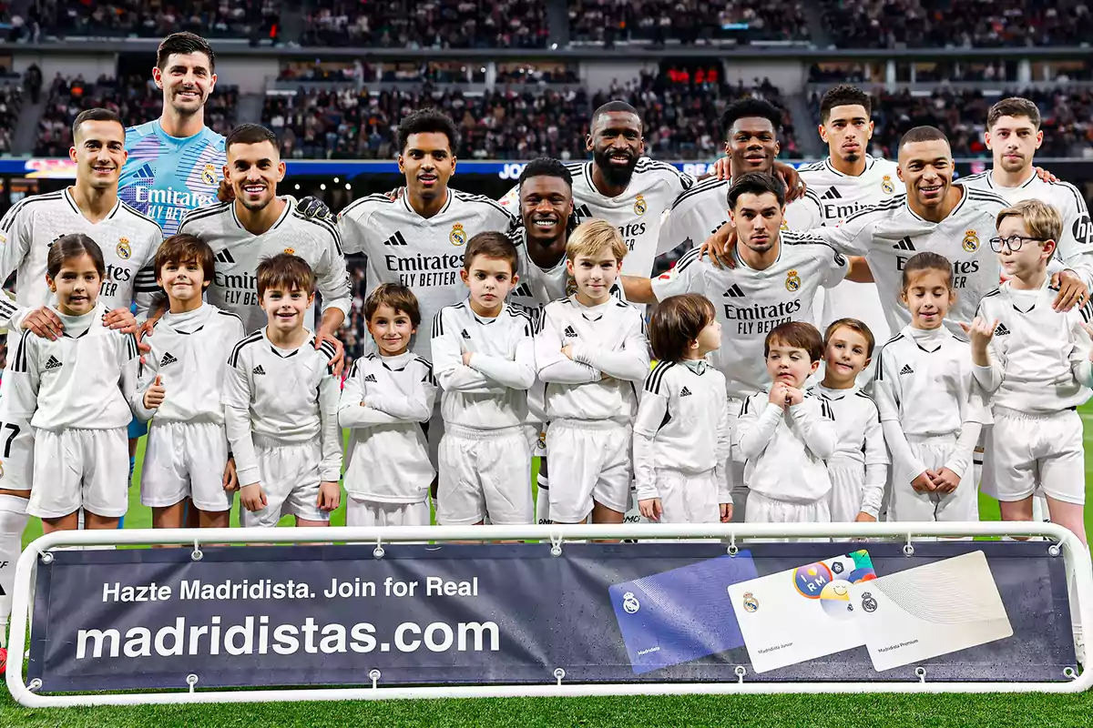 Un grupo de jugadores de fútbol con niños en uniforme blanco posan juntos en un estadio, con un cartel promocional al frente.