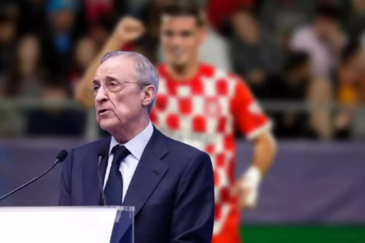 Un hombre mayor hablando en un podio con un fondo desenfocado de un jugador de fútbol con uniforme a cuadros rojos y blancos.