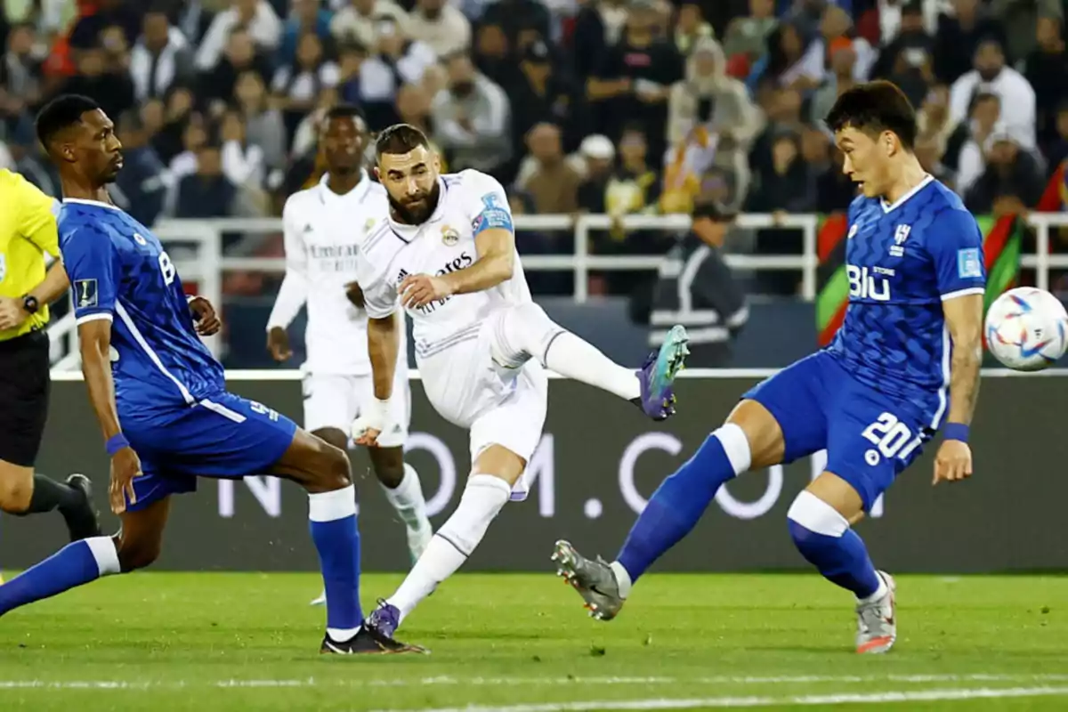 Jugador de fútbol con uniforme blanco patea el balón mientras dos rivales con uniforme azul intentan bloquearlo durante un partido con público de fondo Jugador de fútbol con uniforme blanco patea el balón mientras dos rivales con uniforme azul intentan bloquearlo durante un partido con público de fondo