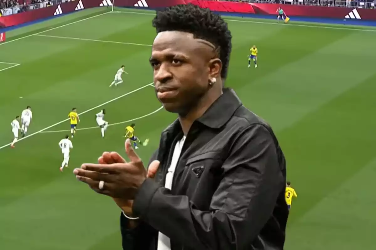 A man applauding with a soccer field in the background where a match is being played between teams with white and yellow uniforms.