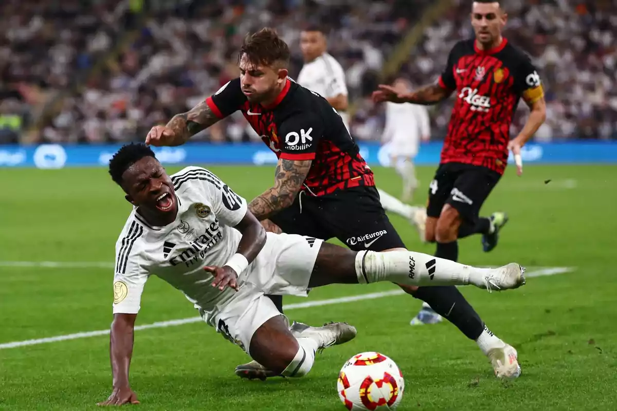 A soccer player in a white uniform falls to the ground as a player in a black and red uniform challenges him during a match. A soccer player in a white uniform falls to the ground as a player in a black and red uniform challenges him during a match.