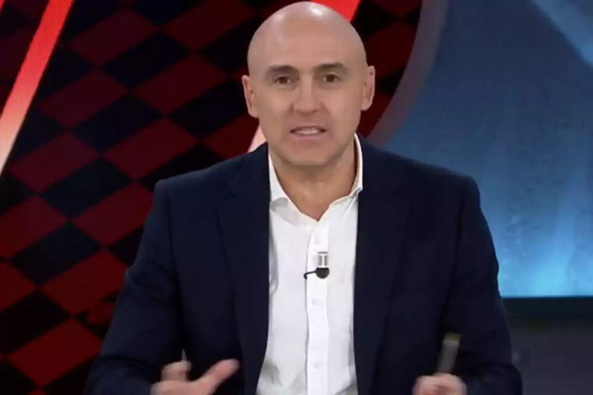 A bald man in a dark suit and white jersey is speaking in a television studio with a geometric design background.