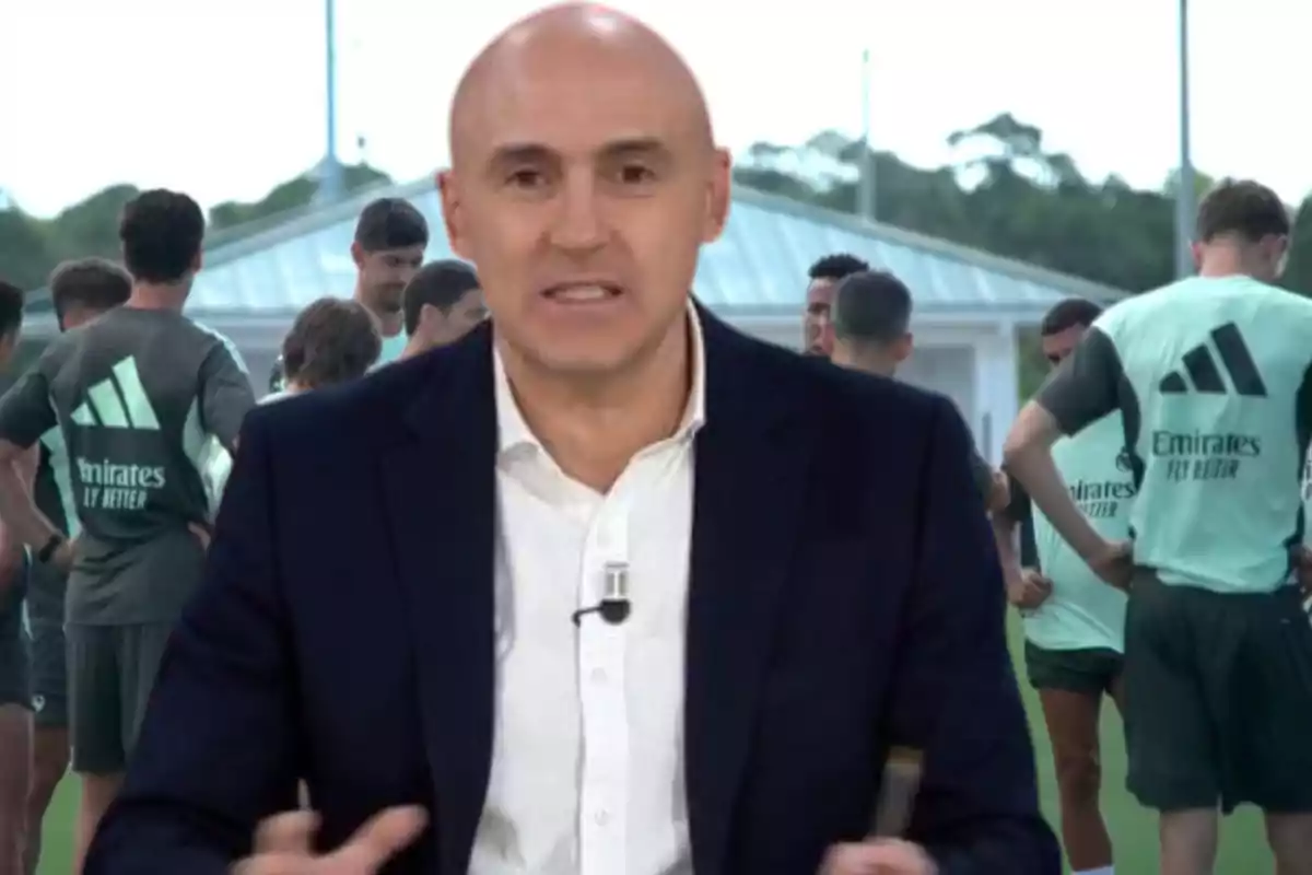 A bald man in a dark jacket and white jersey speaks in front of a group of players wearing green training uniforms with the Emirates Fly Better logo on the back.