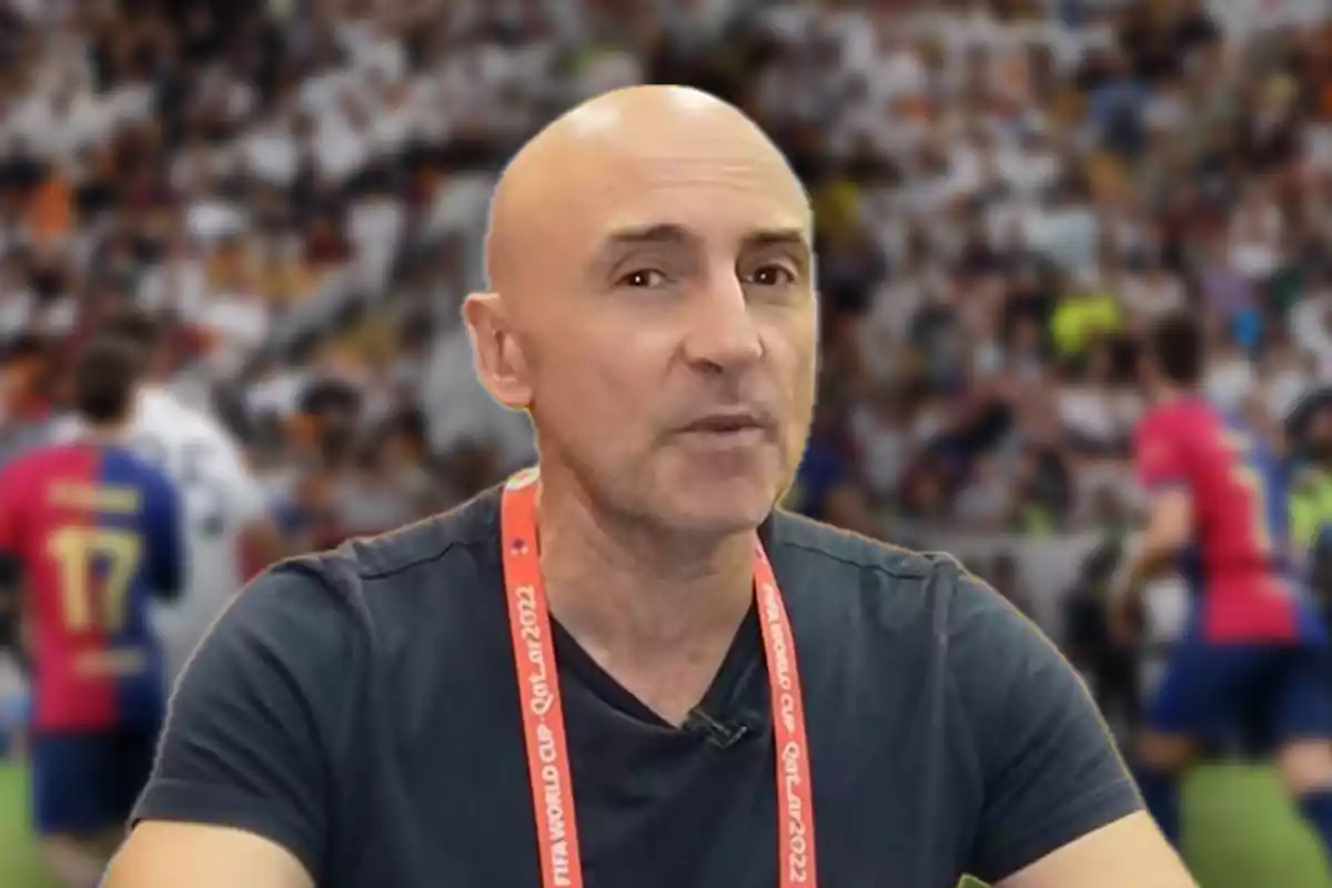 A bald man wearing a dark T-jersey and a 2022 FIFA World Cup badge, with a crowded stadium and out-of-focus football players in the background.