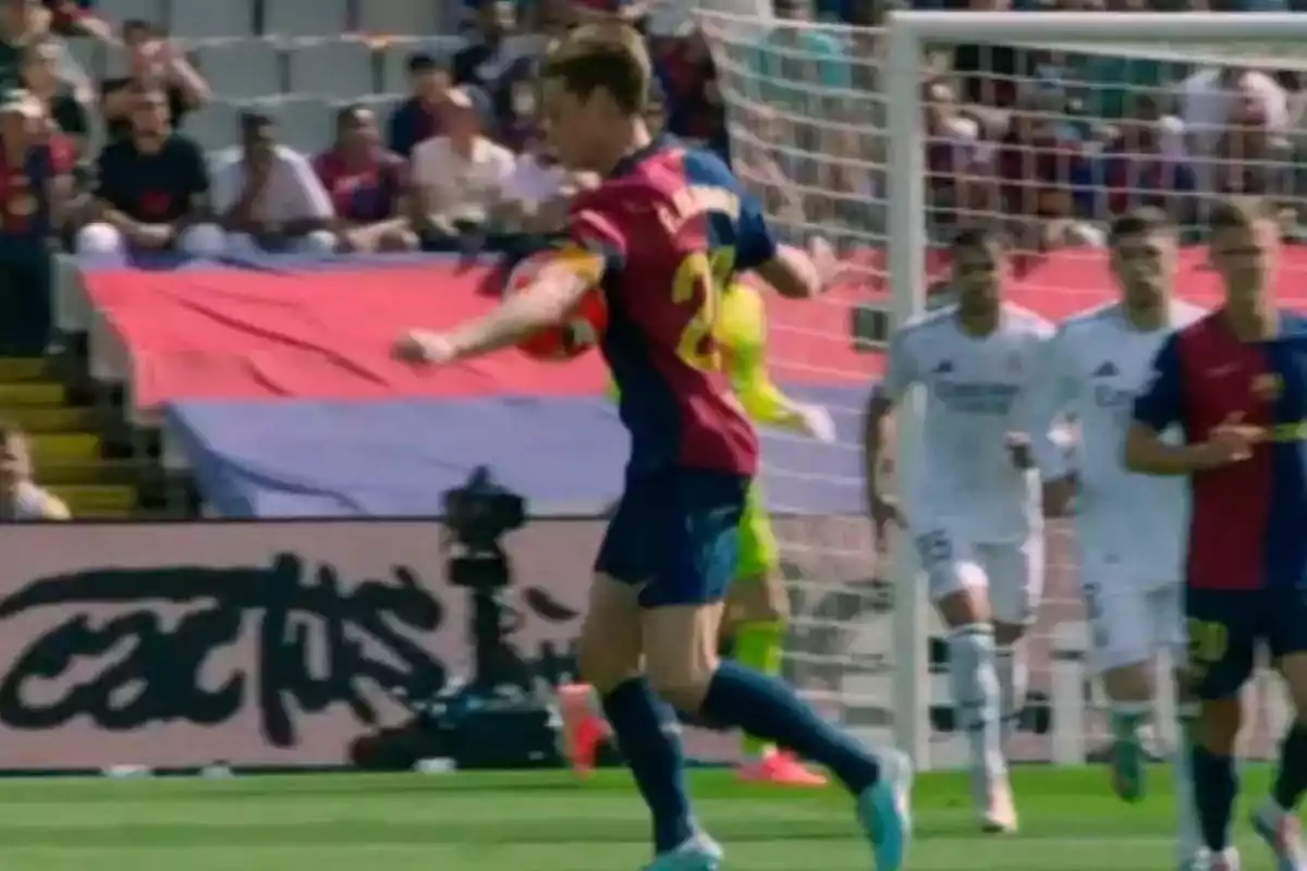 Un jugador de fútbol con uniforme azul y rojo controla el balón en un partido, mientras otros jugadores con uniforme blanco observan cerca de la portería.
