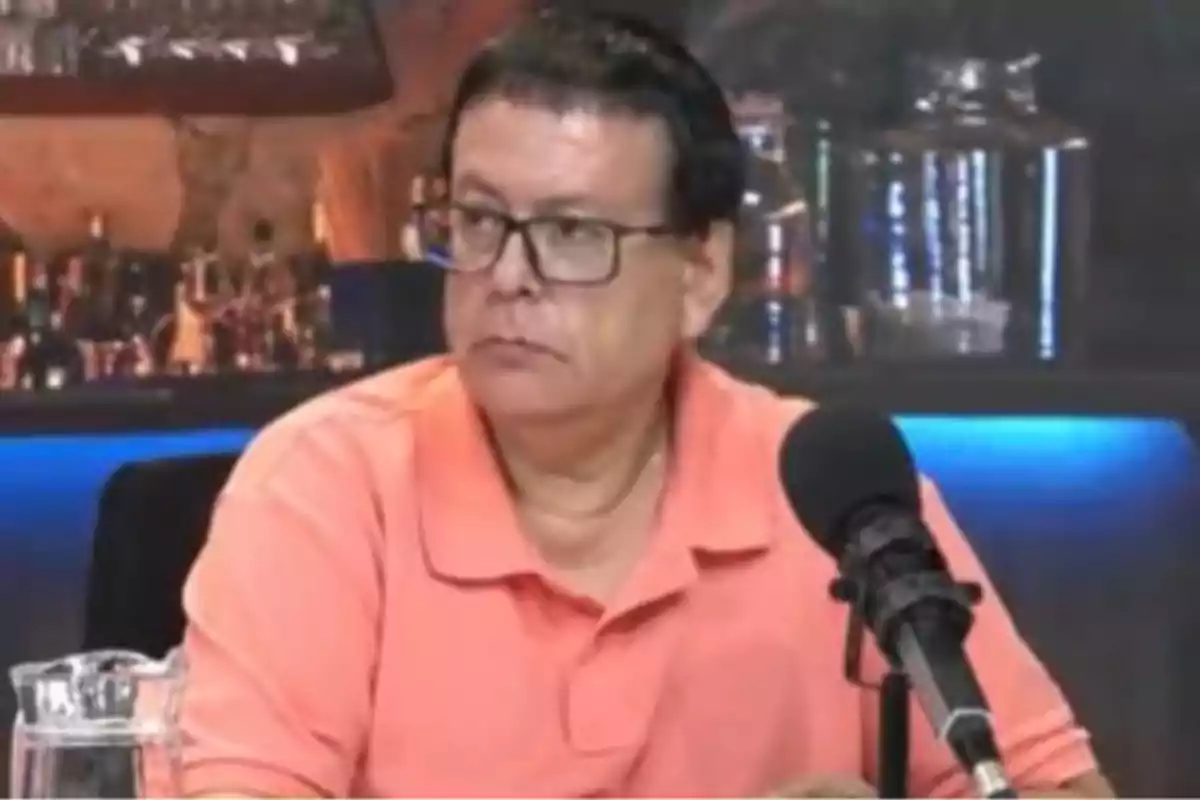 A man with glasses and an orange jersey sits in front of a microphone in a studio with a bar in the background. A man with glasses and an orange jersey sits in front of a microphone in a studio with a bar in the background.