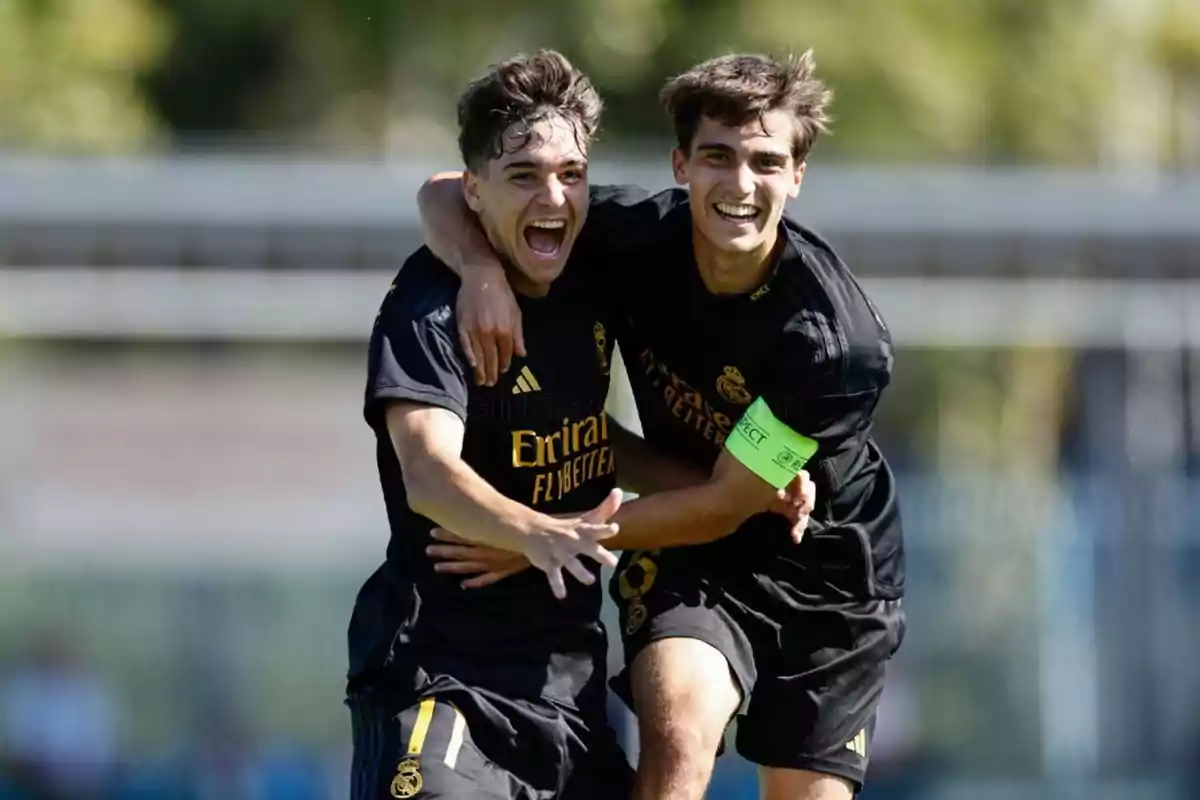Two players celebrate a goal by hugging and smiling, both wearing black uniforms with gold details, and one is wearing a green armband. Two players celebrate a goal by hugging and smiling, both wearing black uniforms with gold details, and one is wearing a green armband.