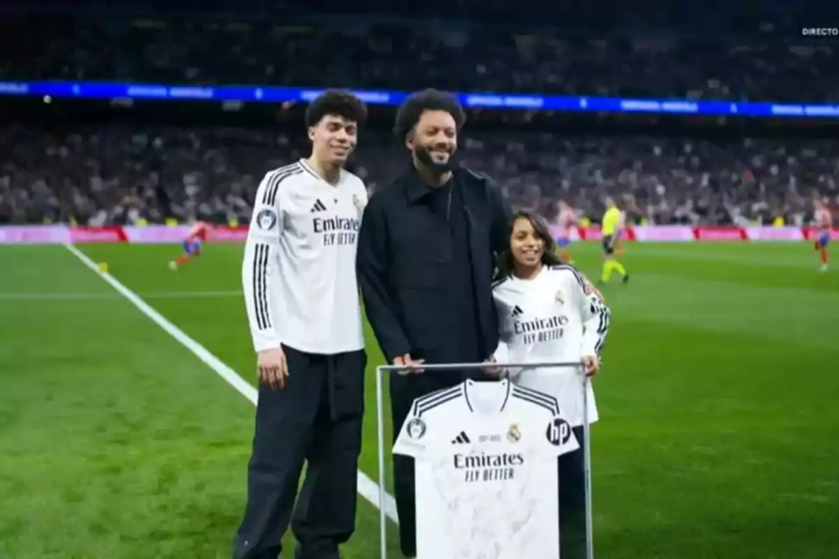 Three people pose on a soccer field with a framed Real Madrid jersey.