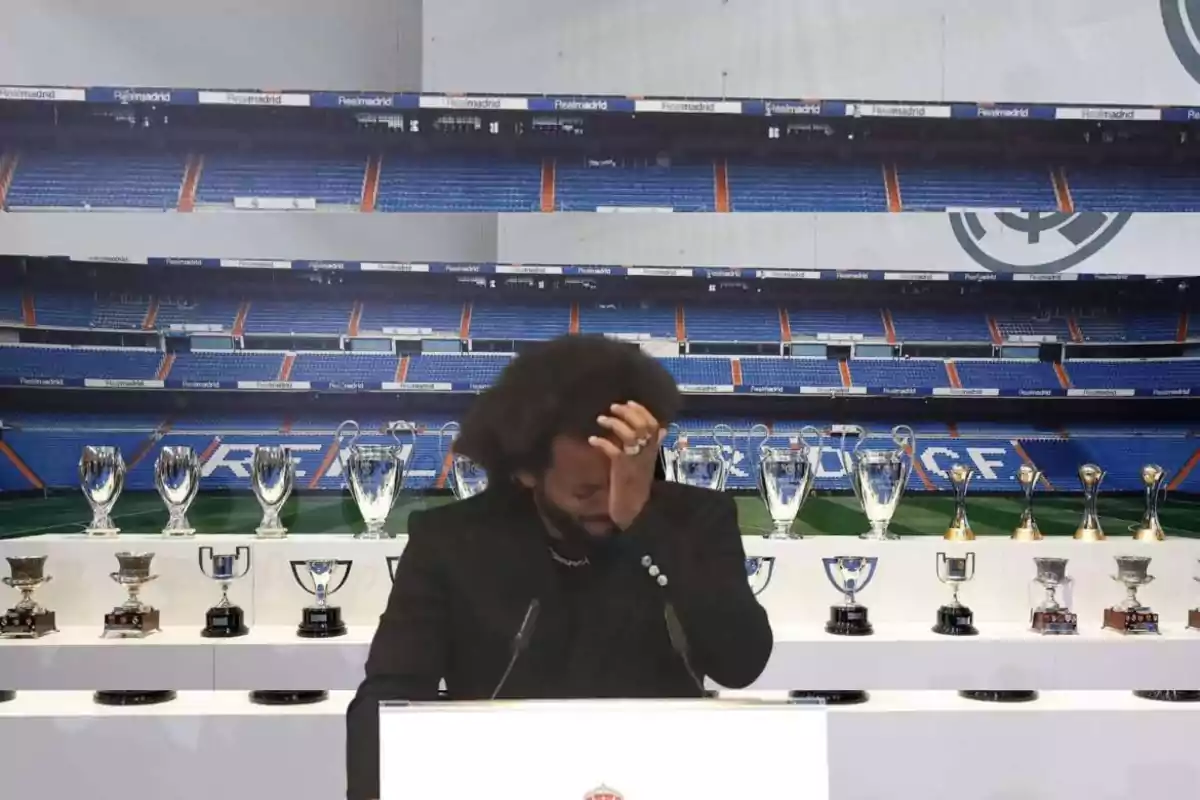 A man in a black suit stands in front of a podium in a football stadium, surrounded by numerous trophies.