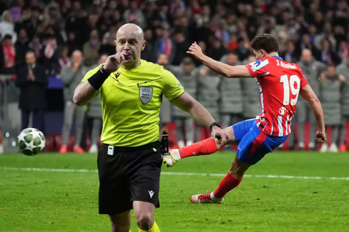 A soccer referee on the field makes a silence gesture while a player from the team in red and white uniform kicks the ball.