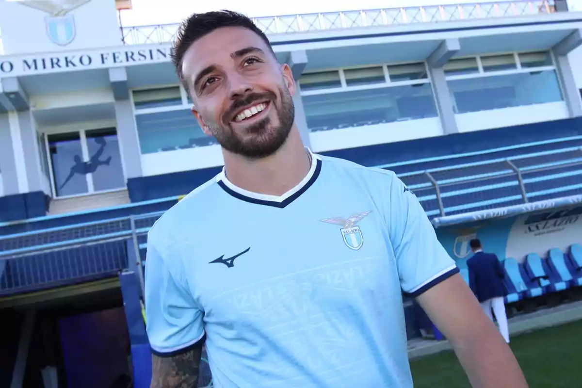 A bearded player wearing Lazio’s light blue jersey smiles in a stadium in front of the stands and the bench area. A bearded player wearing Lazio’s light blue jersey smiles in a stadium in front of the stands and the bench area.
