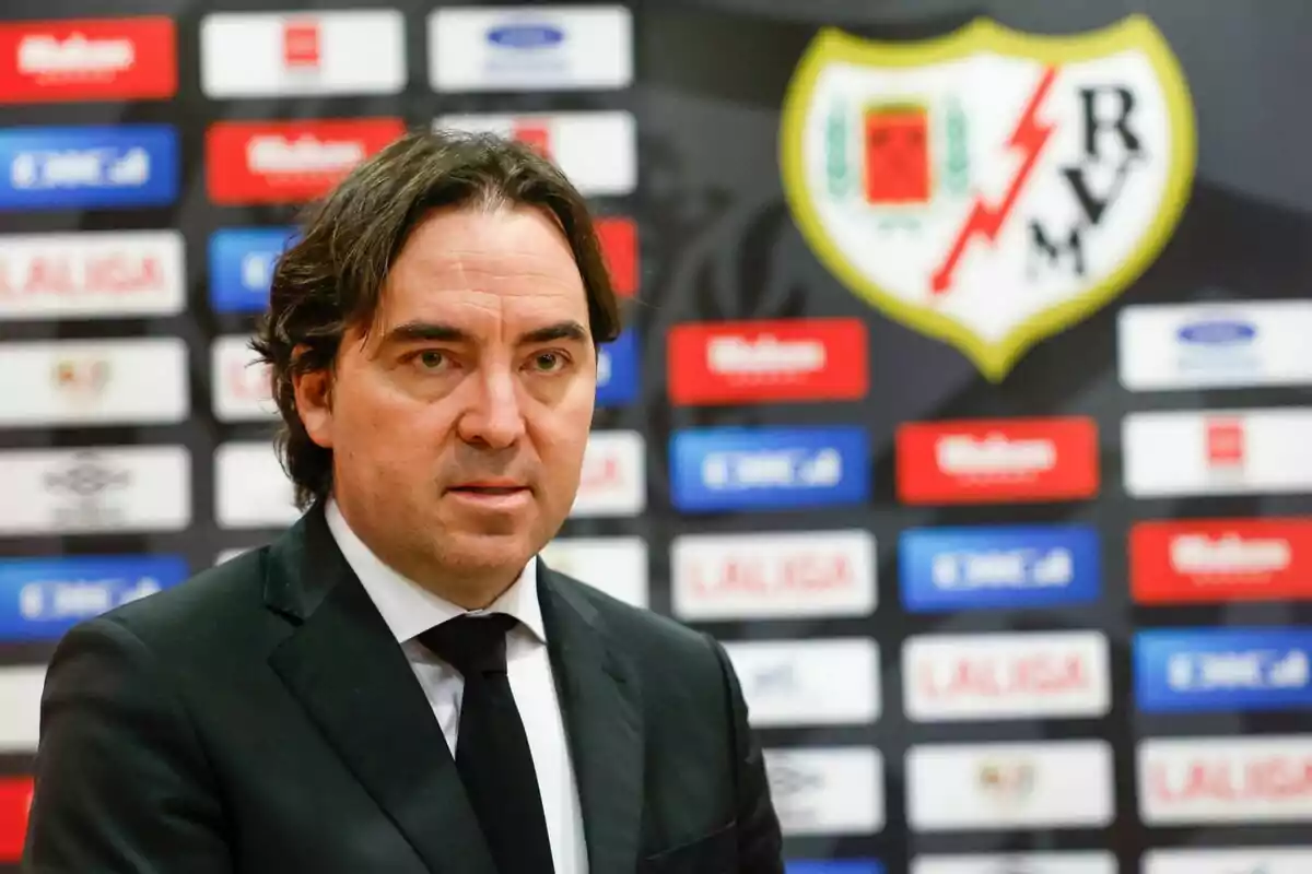 Un hombre con traje oscuro está de pie frente a un fondo con logotipos y el escudo del Rayo Vallecano.