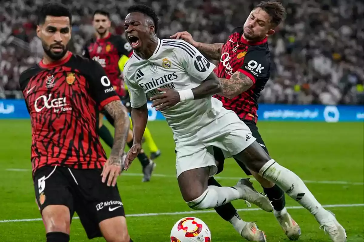 Un jugador de fútbol con uniforme blanco es derribado por un oponente con uniforme rojo y negro durante un partido.