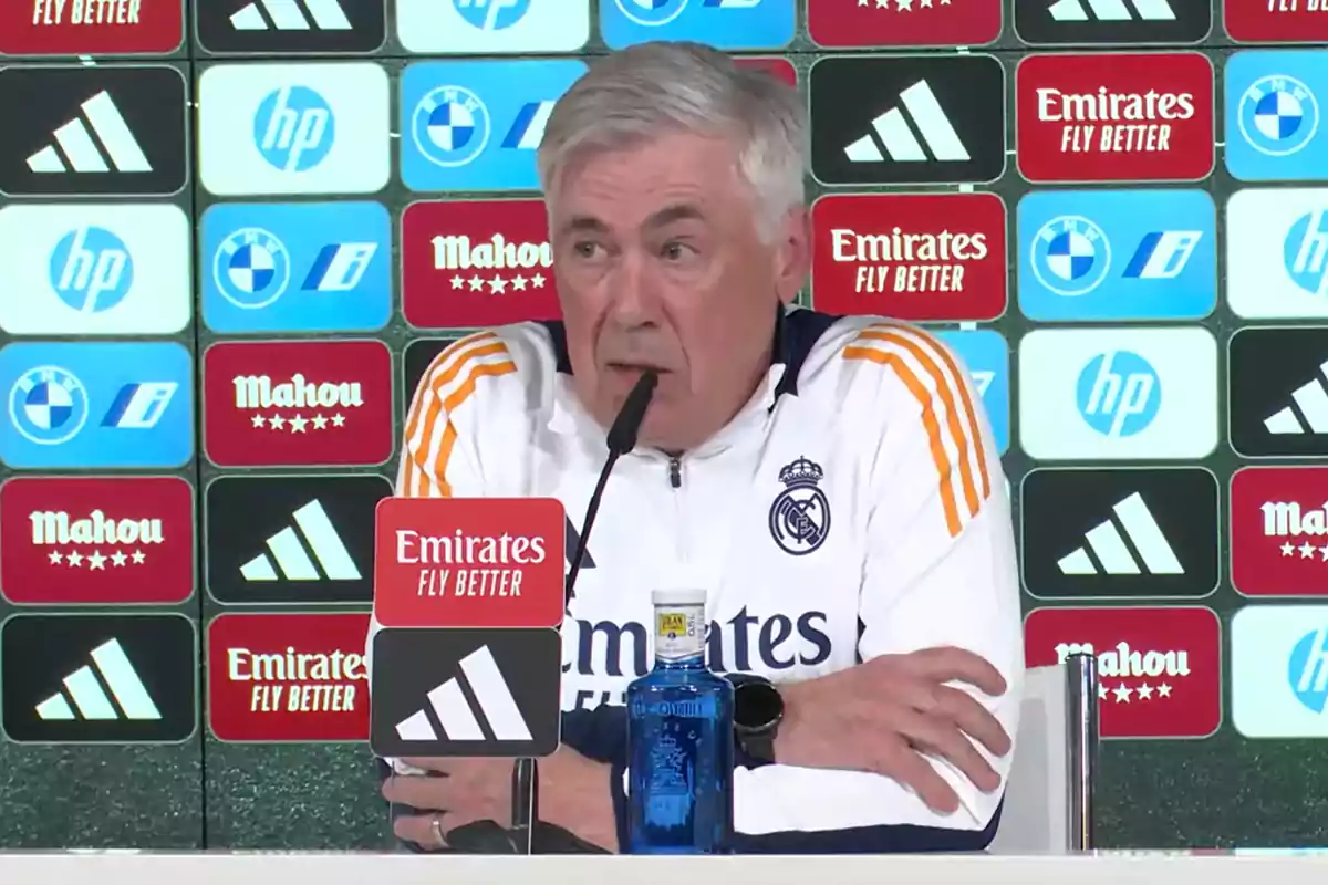 Un hombre en una conferencia de prensa con un fondo de logotipos de patrocinadores deportivos.