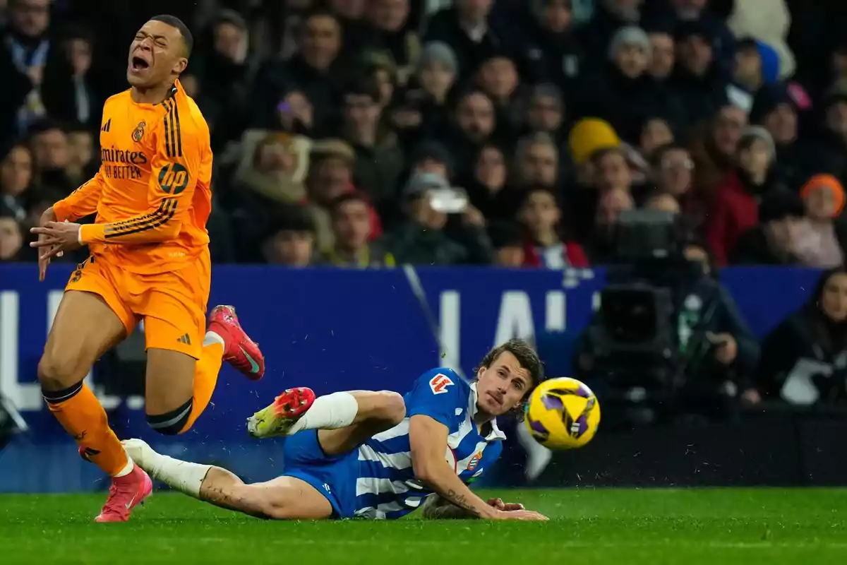 Un jugador de fútbol con uniforme naranja parece estar en dolor mientras otro jugador con uniforme azul y blanco está en el suelo durante un partido.