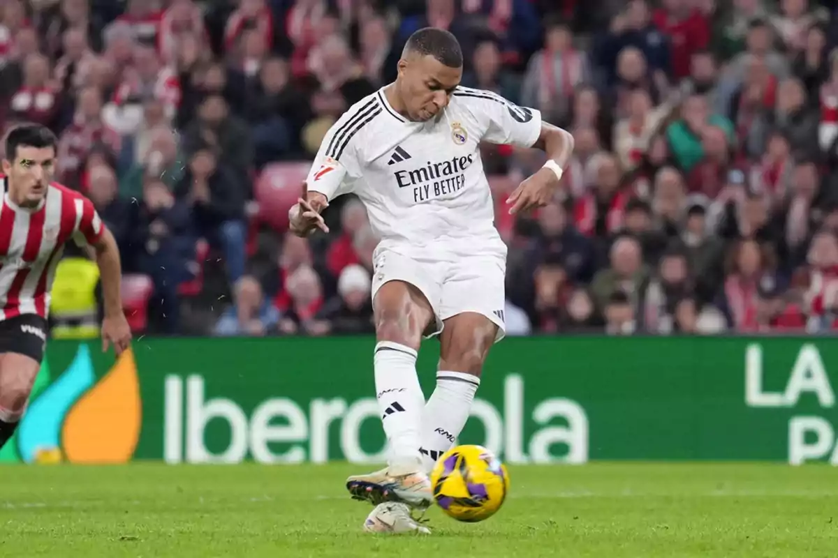 Jugador de fútbol en acción durante un partido con el uniforme blanco del Real Madrid mientras patea el balón en un estadio lleno de espectadores.