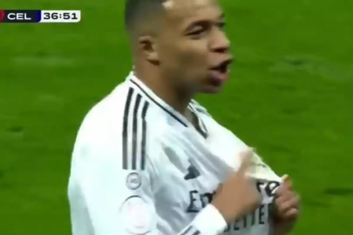 Un jugador de fútbol en el campo con uniforme blanco señalando su camiseta durante un partido.