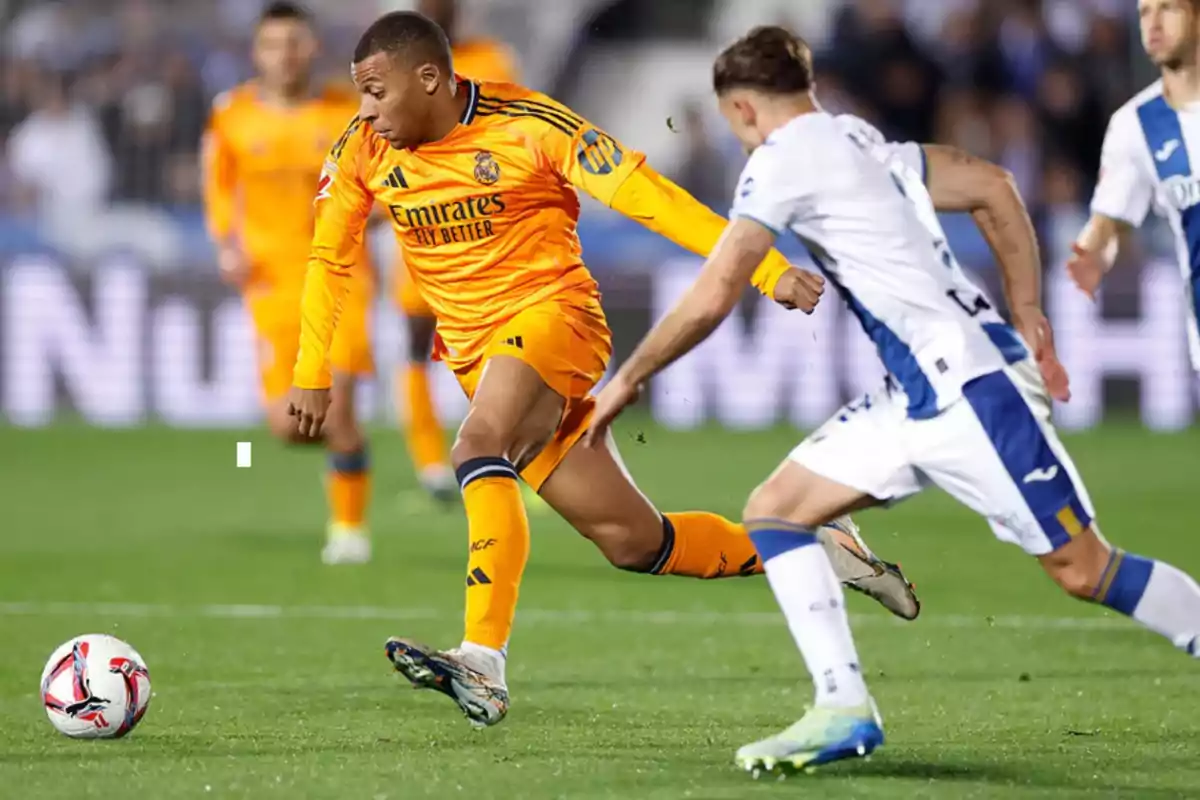 Jugador de fútbol en uniforme naranja controlando el balón durante un partido mientras es perseguido por un oponente en uniforme blanco y azul.