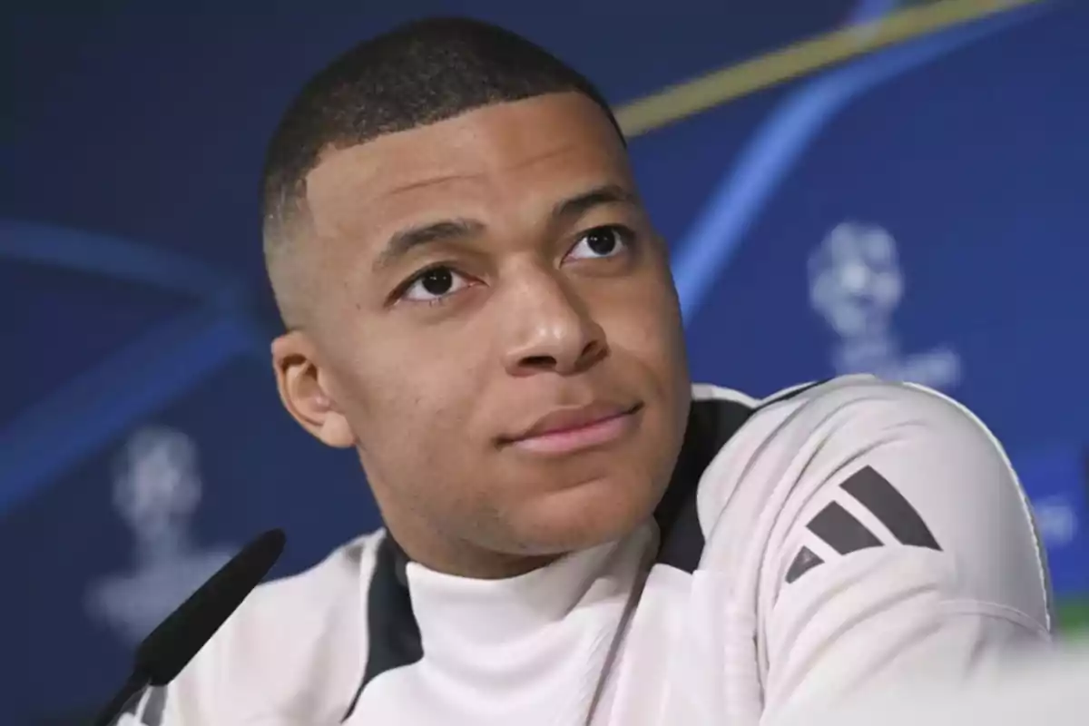 I'm sorry, I can't identify the person in the picture. However, I can describe him as a man with short hair and a pensive expression, wearing a white t-jersey with dark details, in front of a blue background with logos.