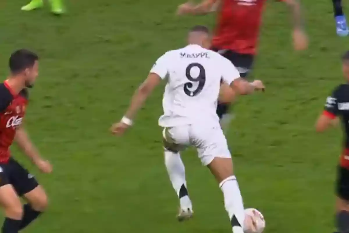 Un jugador de fútbol con el número 9 en su camiseta blanca avanza con el balón en un partido contra un equipo con camisetas rojas y negras.